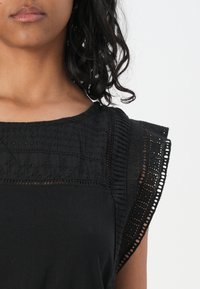 Black sleeveless top featuring textured embroidery on the bodice and delicate lace trim along the shoulder edges. Soft fabric.