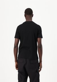 Tricou negru cu mânecă scurtă și guler rotund, cu un design simplu. Un logo discret este imprimat pe gât. Asortat cu pantaloni cargo negri.