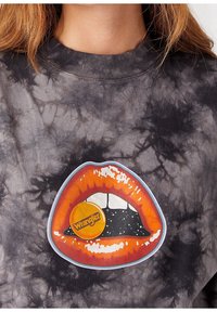 Sudadera negra con efecto tie-dye que presenta un gráfico de una boca abierta con labios naranjas y un logo circular de "Wrangler" en su interior.