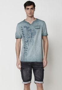 Mėlynas medvilnės T-shirt su v-kaklu, trumpomis rankovėmis ir priekine grafika. Suderinta su tamsiais džinsiniais šortais, turinčiais apvyniotus kraštus ir kišenes.