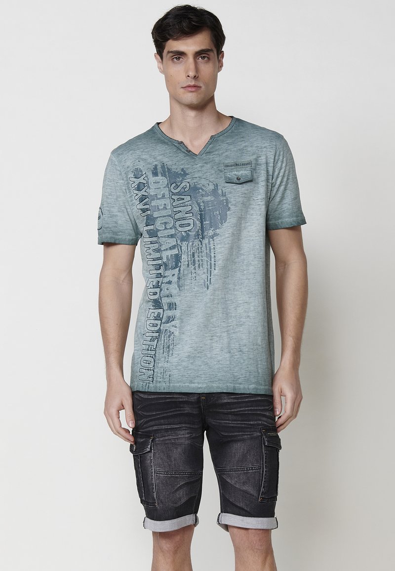 Mėlynas medvilnės T-shirt su v-kaklu, trumpomis rankovėmis ir priekine grafika. Suderinta su tamsiais džinsiniais šortais, turinčiais apvyniotus kraštus ir kišenes.