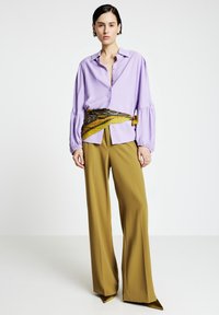 Hellviolette Hemdbluse mit Puffärmeln, kombiniert mit hoch taillierter, weiter olivgrüner Hose. Accessoires: Ein gemusterter Halstuchgürtel.