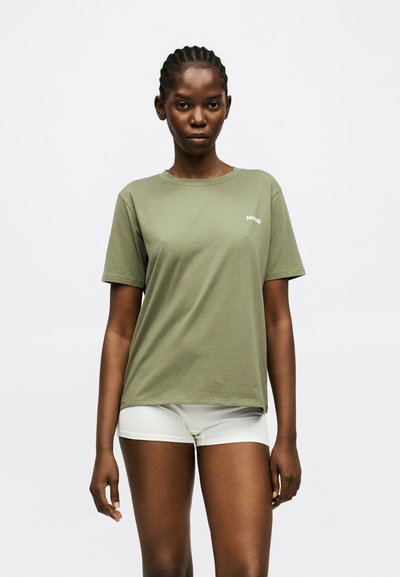 Femme aux cheveux tressés portant une chemise à manches courtes vert olive et un short blanc, debout devant un fond blanc uni.