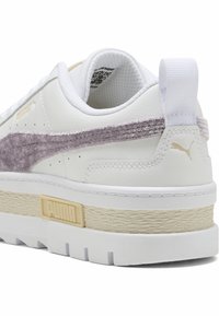 Baltie sporta apavi ar purpura samta svītru, teksturētu bēšu vidusdaļu, zelta akcentu logotipu un polsterētu rotu komfortam.