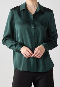 Tummanvihreä satiinipusero, jossa on napitettava etuosa, pitkät hihat ja terävä kaulus, sileä pinta ja rento istuvuus.