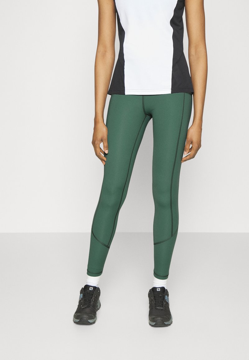 Leggings de sport verts avec un tissu texturé, présentant des coutures noires contrastées et une coupe ajustée, associés à des baskets noires et des chaussettes blanches.