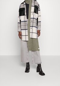 Checked outer jacket in cream, black, and blue, with leather detail. Layered with a green knitted scarf over a black top and gray wide-leg jeans.