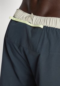 Mörkblå shorts med elastisk midjeband, som har en ljusgrå inre foder och en liten ljusgrön accentöggla baktill.