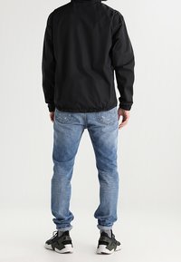 Black, lightweight jacket with elastic cuffs, paired with light blue jeans. Footwear features a chunky design in black and white with green accents.