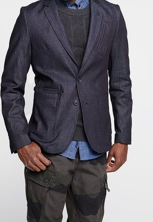 Blazer en denim foncé avec revers en notch, deux poches avant et fermeture à un bouton. Porté sur un pull noir texturé et une chemise bleue.