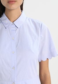 Ljust blå skjorta med krage och knäppning, med scallop-kantade korta ärmar och randig textur.