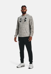 Grau meliertes Hoodie mit schwarzem Logo, weißen Zugbändern; kombiniert mit schwarzen Jogginghosen und weißen Sportschuhen mit grauen Akzenten.