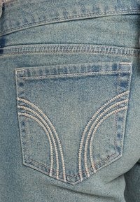 Υφασμα από ανοιχτό μπλε τζιν με πίσω τσέπη και λευκή ραφή σε καμπύλο μοτίβο. Η υφή είναι λεία με κλασική εμφάνιση τζιν.