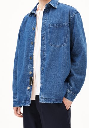 Persona che indossa una giacca di jeans blu sbottonata sopra una camicia chiara con una mano nella tasca dei pantaloni scuri.