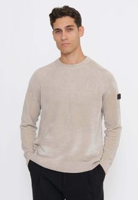 Beiger Strickpullover mit gerippten Schulternähten und rundem Ausschnitt. Verfügt über ein kleines schwarzes Logo auf dem linken Ärmel. Weiche Textur.