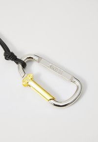 Moschettone argento con cordone in pelle nera, caratterizzato da un design a colonne gialle e incisa la parola "ARIES" sul lato.