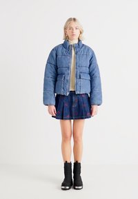 Denim puffer jacket in light blue, cropped design with front pockets, worn over a beige sweater. Paired with a pleated denim skirt and black boots.