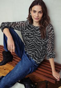Blusa trasparente grigia e nera con motivo a zebra, scollatura rotonda e maniche lunghe gonfiate, abbinata a jeans in denim blu e stivali neri alla caviglia.