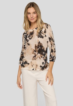 Frau trägt ein beige-schwarz florales Strickoberteil mit Dreiviertelärmeln und hellbeige, weit geschnittene Hosen.