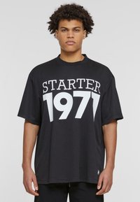 Crna oversize majica s velikim bijelim printom "STARTER 1971" na prednjem dijelu. Mekani materijal s okruglim izrezom i kratkim rukavima.