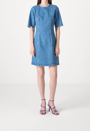 Kvinde iført en knælange blå denimkjole med frynsede kanter og korte ærmer, kombineret med lilla sandaler med snørre og høje hæle.