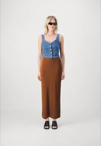 Haut sans manches en denim avec des boutons sur le devant, associé à une jupe marron côtelée longeant les chevilles. Des sandales noires à enfiler complètent le look.