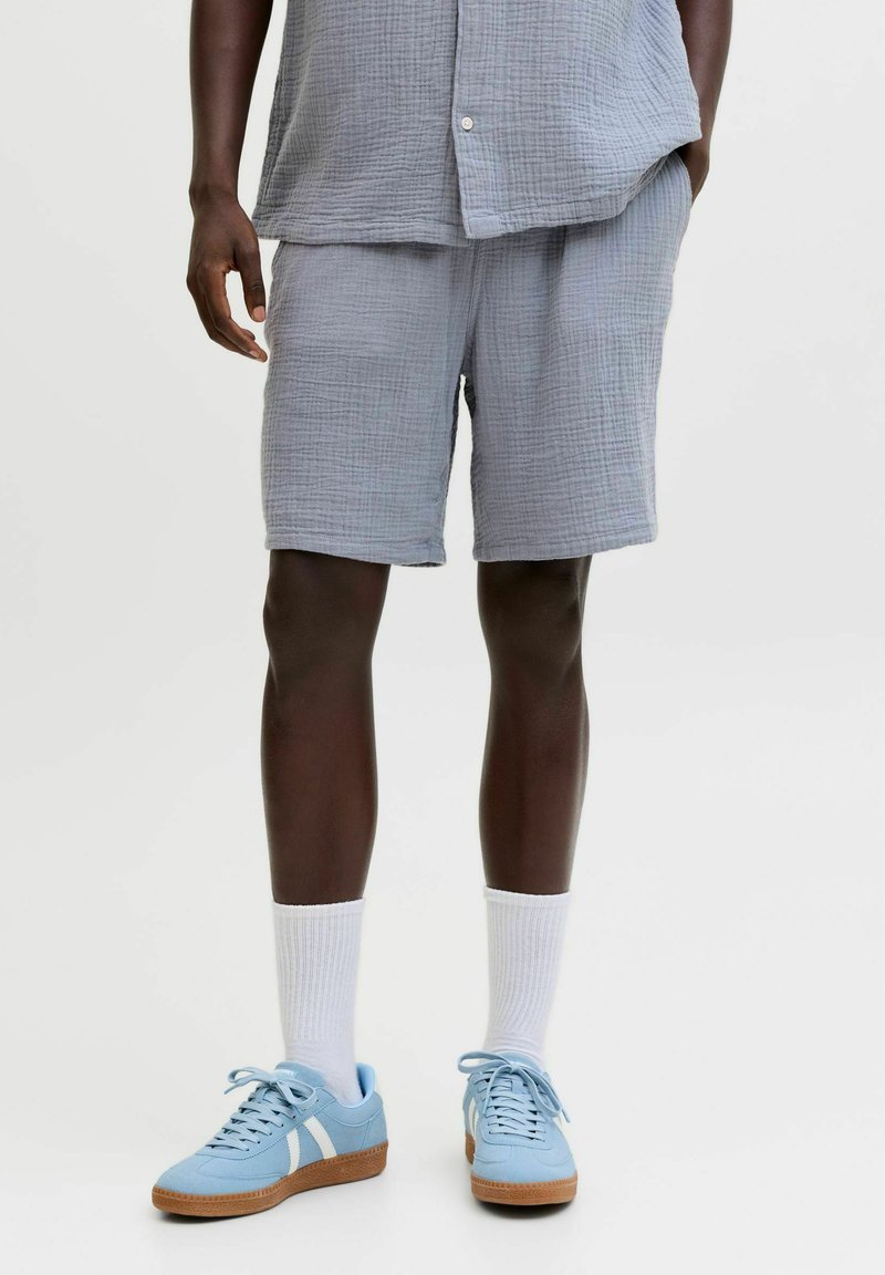 Homme portant un short gris clair texturé et une chemise assortie, des chaussettes blanches montantes et des baskets bleu clair avec semelles en gomme, debout devant un fond uni.