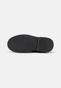 Black shoe sole made of textured rubber, featuring a reinforced heel with gold-colored rivets, flat and slightly rounded shape.