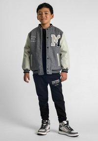 Sivý varsity blejzer s krémovými rukávmi, obsahuje nášivku 'M' a vyšívanie. Čierne nohavice s bielym textom, skombinované so sivými vysokými teniskami.