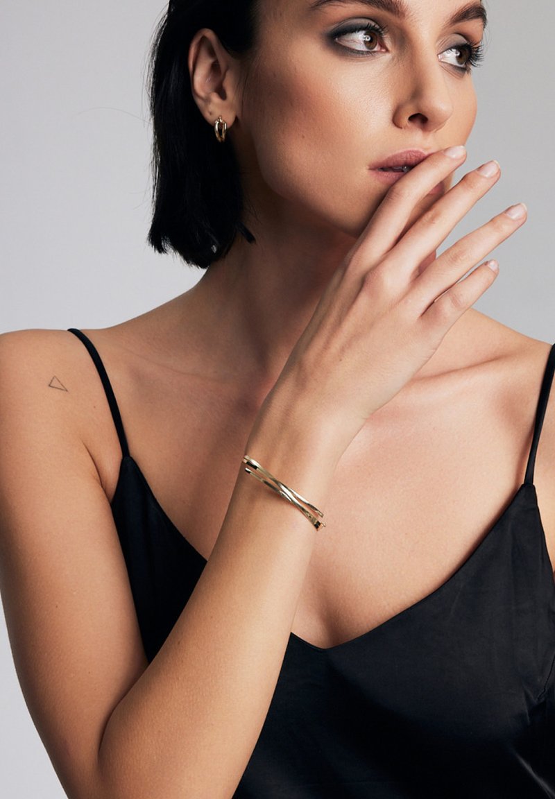 Bransoletka złotego koloru z skręconym wzorem, gładką teksturą, noszona na nadgarstku, w zestawieniu z czarną satynową koszulką na ramiączkach.