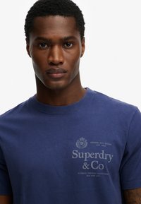 Camiseta de algodón azul marino con cuello redondo. Presenta una impresión en gris claro del logo y texto "Superdry & Co" en la parte delantera. Mangas cortas.