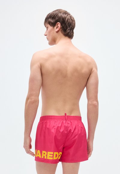 Κόκκινα μαγιό βερμούδα από ελαφρύ ύφασμα, με ελαστική μέση και μεγάλο κίτρινο τύπωμα "ARE D?" στην πίσω πλευρά.
