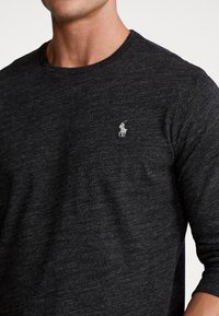 Dunkelgraues Langarmshirt aus strukturiertem Material, mit einem kleinen weißen Logo auf der linken Brust und einem Rundhalsausschnitt.