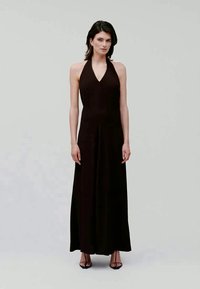 Ženska stoji oblečena v brezrokavni črni halter maxi obleki in črnih sandalih s tankimi petami na enobarvni sivi ozadju.