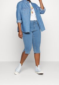 Camicia di jeans, azzurro chiaro; t-shirt bianca con stampa floreale; pantaloni corti fino al ginocchio, azzurro chiaro; sneakers alte azzurre con dettagli a occhiello.