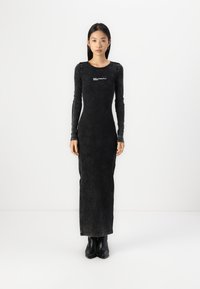 Svart, långärmad klänning med en figurnära silhuett, texturerat tyg och en logodetalj på bröstet. Buren med svarta ankeldöljande stövlar.