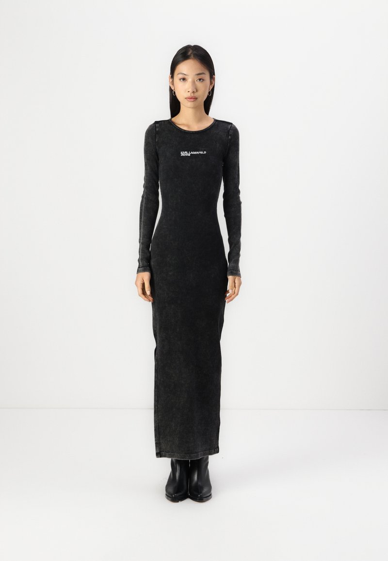 Svart, långärmad klänning med en figurnära silhuett, texturerat tyg och en logodetalj på bröstet. Buren med svarta ankeldöljande stövlar.