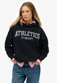 Mujer con las manos en los bolsillos, vistiendo una sudadera negra con el texto "ATHLETICS SD.UNIV.DPT", una camiseta a rayas rosas debajo y jeans azules, contra un fondo blanco.