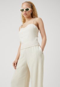 Camisole en lin beige clair avec des bretelles fines, associée à un pantalon large plissé. Le mannequin porte des lunettes de soleil oversize vert clair.
