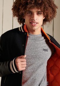 Zwarte en oranje varsityjack met gestreepte boorden, over een grijze T-shirt met een logo, met een gewatteerde binnenkant.