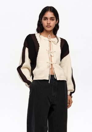 Mujer vestida con un cárdigan de punto en color crema y marrón oscuro con tres lazos delanteros, y pantalones negros de talle alto y pierna ancha, sobre un fondo blanco.