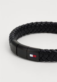Bracelet en cuir tressé noir avec un fermoir noir présentant un petit logo rectangulaire rouge, blanc et bleu.