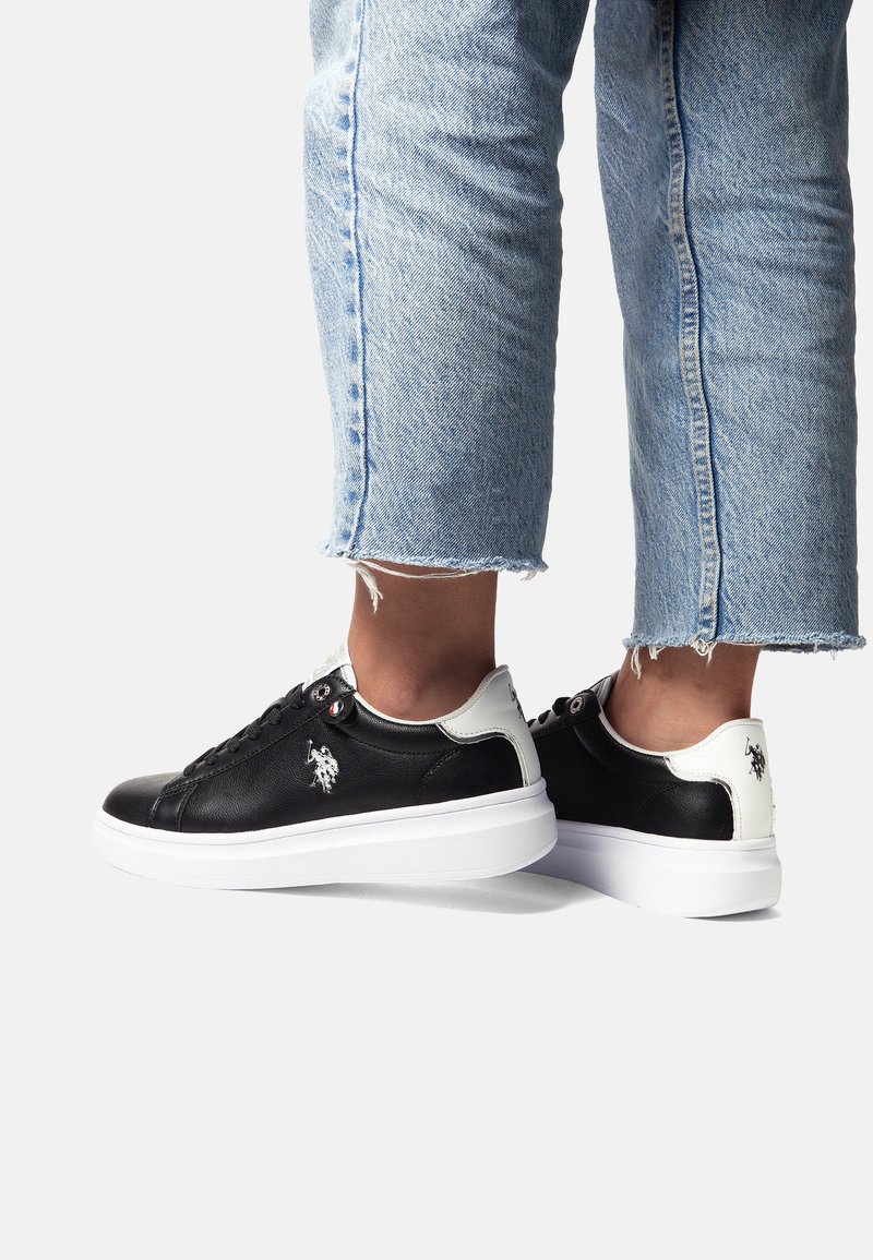 Black leather sneakers with white sole, featuring a padded collar and logo detail. Jeans are cuffed at the ankles, exposing the shoes.