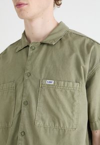 Chemise à manches courtes olive, à boutons sur le devant, avec deux grandes poches, légèrement brillante, avec l'étiquette "Lee" sur la poche. Confectionnée en mélange de coton.