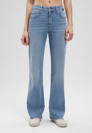 Model wearing light blue flared jeans and white sneakers, standing against a plain gray background.