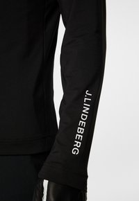 Musta pitkähihainen paita, jossa on sileä pinta ja valkoinen logo hihassa. Suunnittelu on istuva ja siinä on litteät saumat.