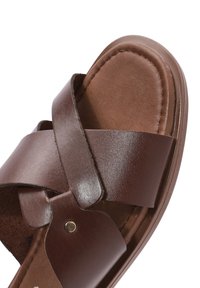 Sandale en cuir marron avec des brides entrecroisées, semelle plate et semelle texturée. Présente un accent métallique près de la bride.