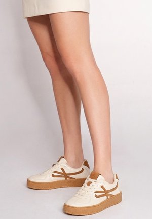 Gambe con sneakers bianche e marroni con lacci e suole spesse in gomma, in piedi su uno sfondo chiaro e uniforme.