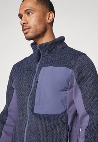Veste en polaire avec un col montant, en tissu texturé bleu marine, avec des accents lisses et une poche poitrine zippée dans une teinte violette contrastante.