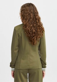Veste vert olive à coupe ajustée, manches longues et poignets rayés. Tissu texturé et coutures cintrées pour un look structuré.